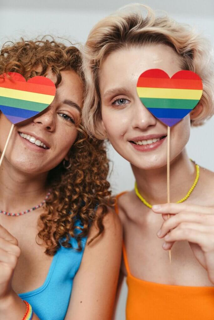 λεσβίες που κρατούν από ένα σημαιάκι της LGBTQ+ κοινότητας σε σχήμα καρδιά μπροστά από το ένα τους μάτι
