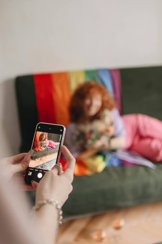 woman hands taking photo of a lesbian woman or queer person sitting on a sofa with the rainbow flag