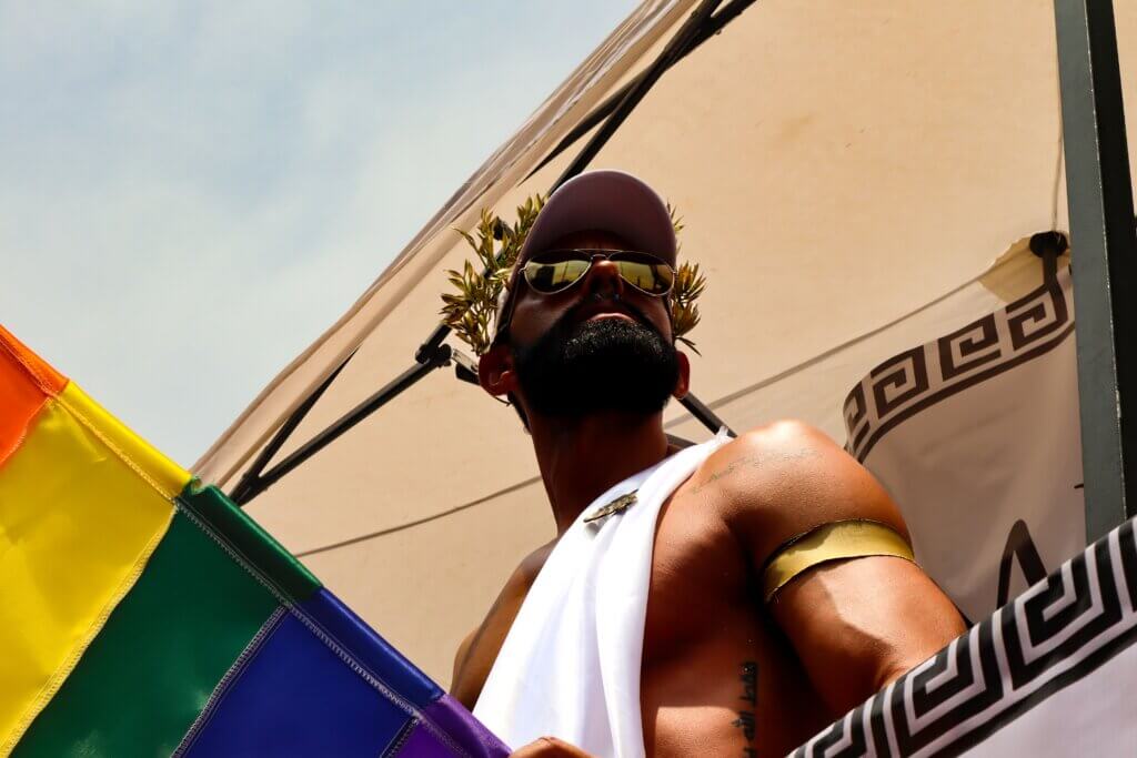 LGBTQ+ άνδρας ντυμένος ως αρχαίος Έλληνας θεός που κρατάει την πολύχρωμη σημαία