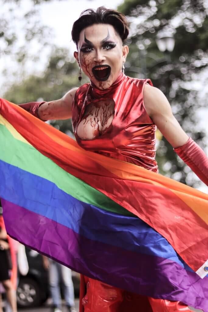 trans person with blood on their chest keeping the LGBTQ+ flag and screaming